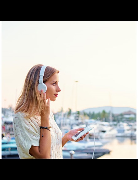 An image showing a woman in a marina. She has an iPhone in one hand and large white headphones on her head. She has her other hand raised to one side of the headphones. There are boats moored in the background.
