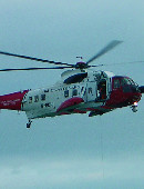 Photograph showing a red and white helicopter in flight against a grey sky. The photo is taken from the helicopter's starboard side. The door is open and a person in orange overalls can be seen standing inside. There are seven windows along the starboard side. Text on the helicopter reads, \"Coastguard rescue\", \"HM Coastguard\", and \"G-B01J\".