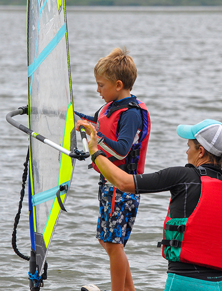 Young boy stood on windsurfing board holding sail. Female instructor stood behind him, helping. 